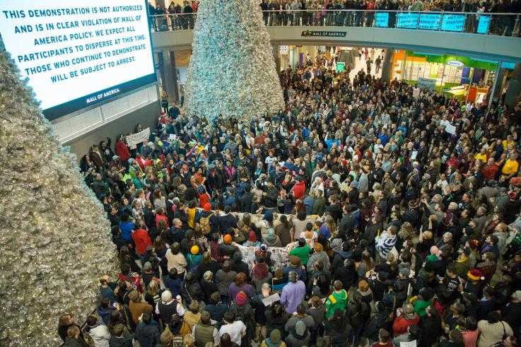 A huge group of protesters stand under an electronic sign in a shopping mall during a protest. The sign demands they disperse or face arrest.