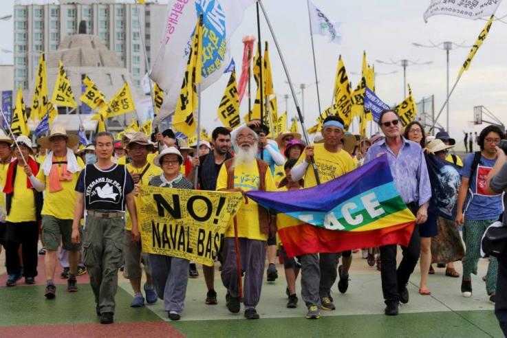 A large number of people march towards the camera. They are wearing yellow and carrying yellow flags reading "No Naval base"
