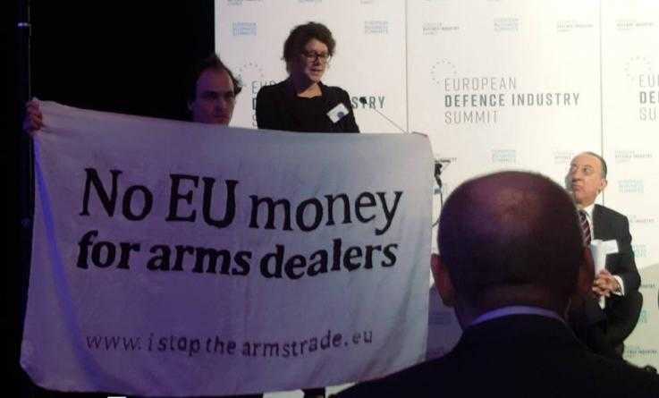 An activist stands on a stage at the microphone. Another holds a banner reading "No EU monday for arms dealers." A man in a suit on the stage looks on.