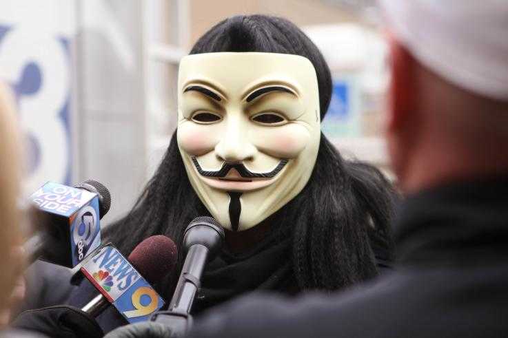 An activist wearing a guy fawkes mask speaks to the media