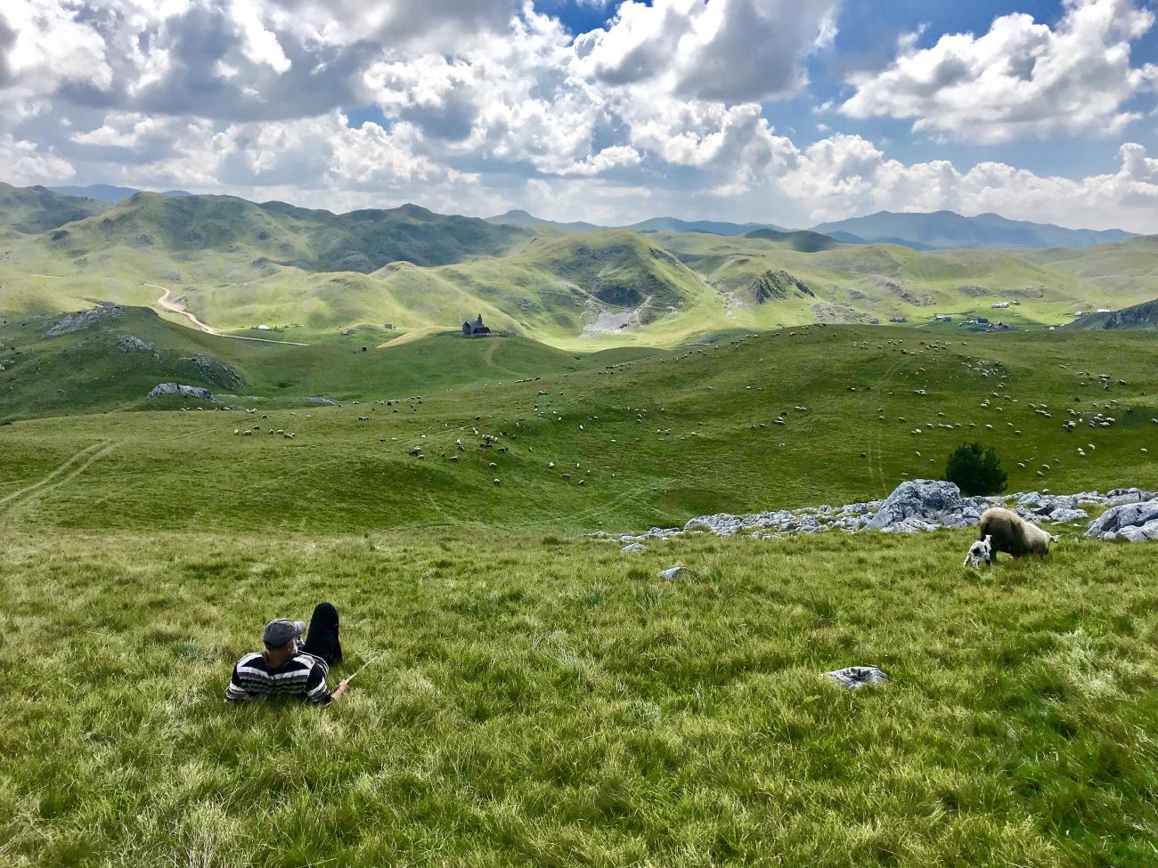 The Sinjajevina highlands - rolling green hills. In the background is a small church. In the foreground a man relaxes on the grass. To the right of the photo there is a sheep and lamb.