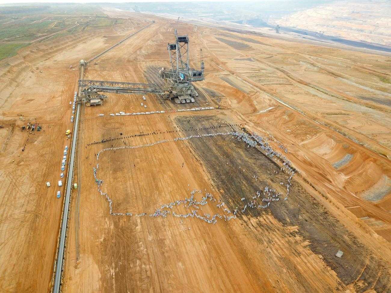 A huge barren landscape with yellow soil. In the background is a huge mining machine. In the foreground but still tiny are hundreds of people dressed in white, forming a circle