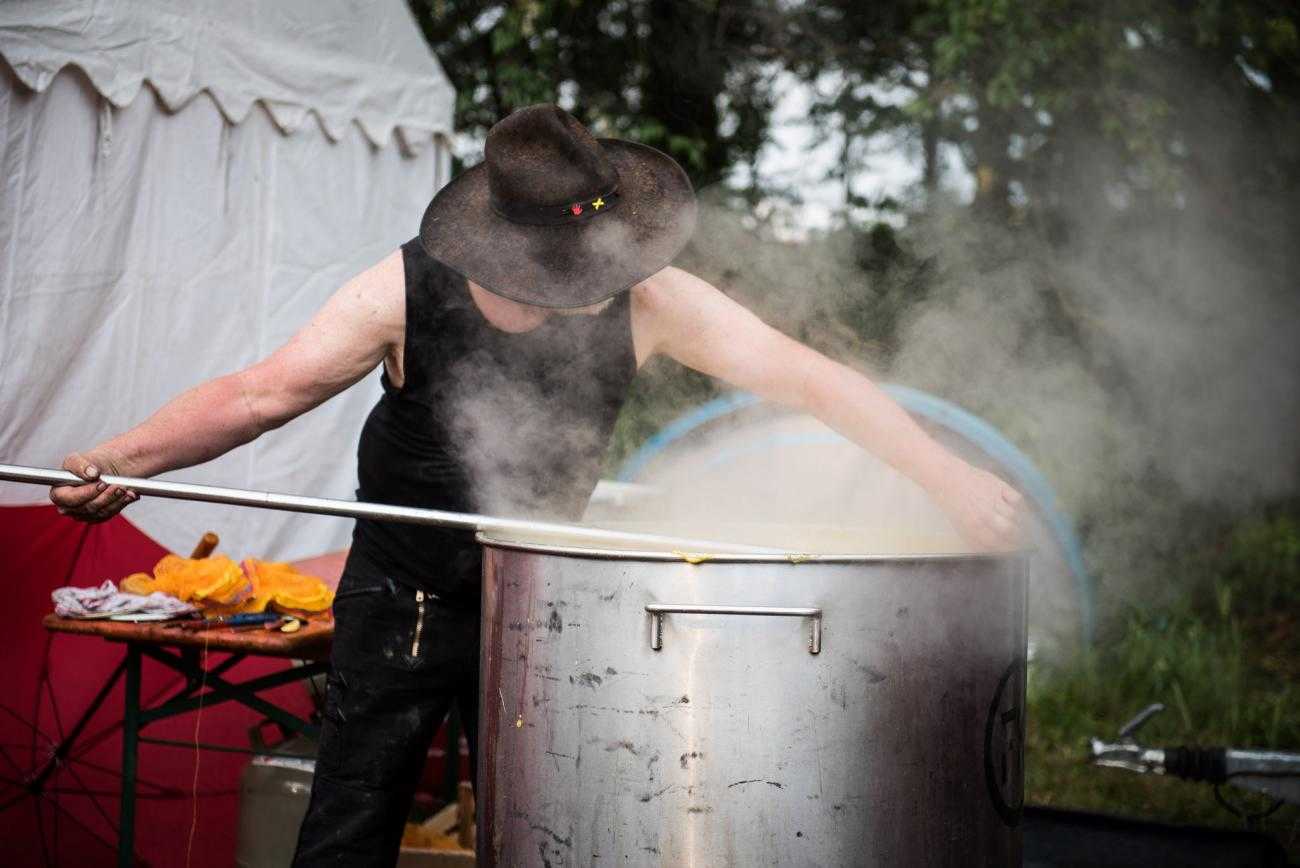 Someone cooks over a large pot. Their face is obscured with a large hat