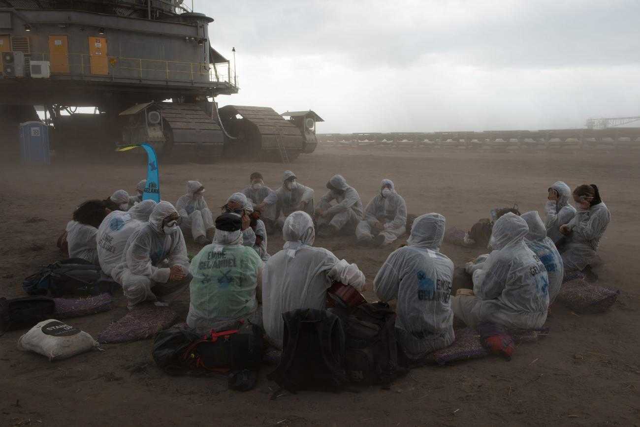A dozen activists sit on the ground in a dusty coal mine. In the background is a huge piece of mining machinery