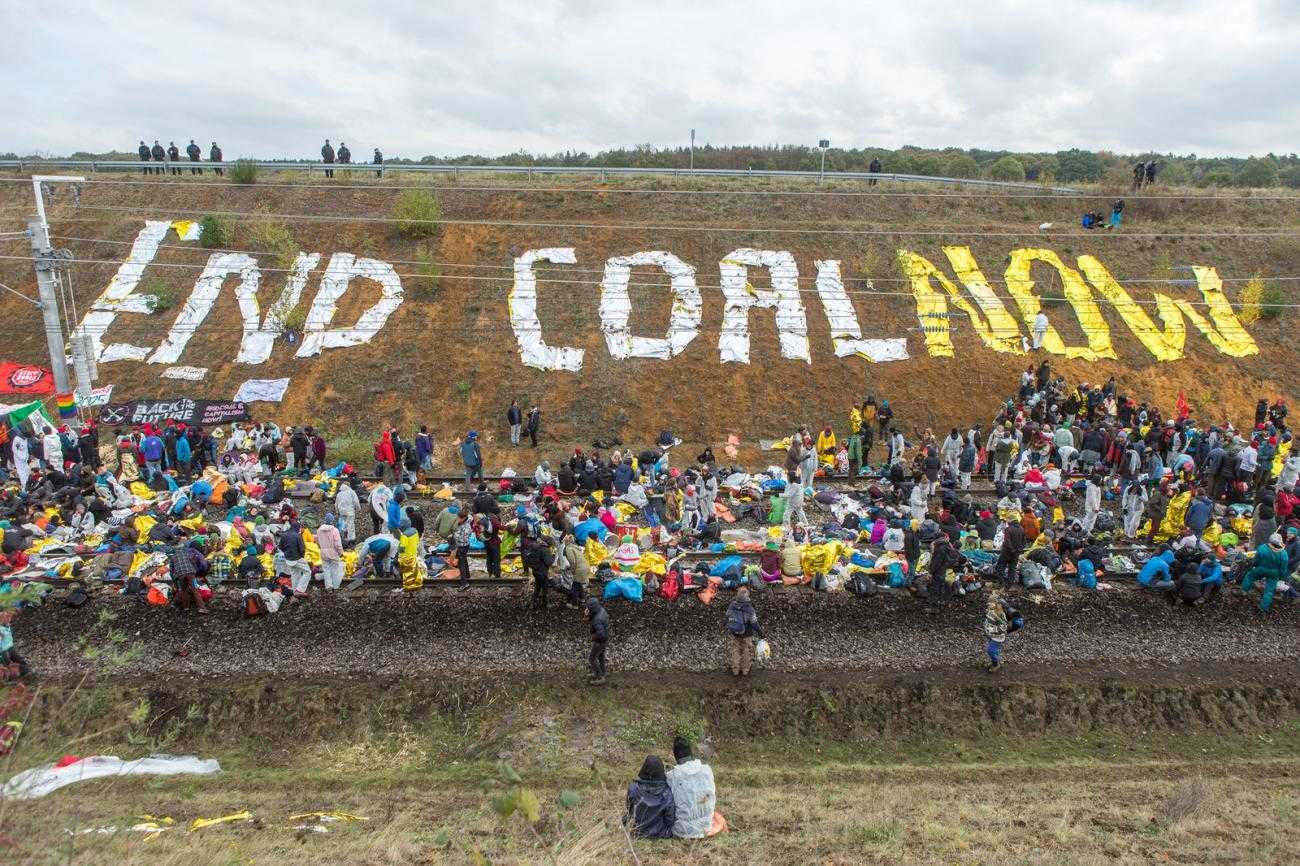A large crowd sits on a railway track. On the bank the words "End Coal Now" have been spelt out