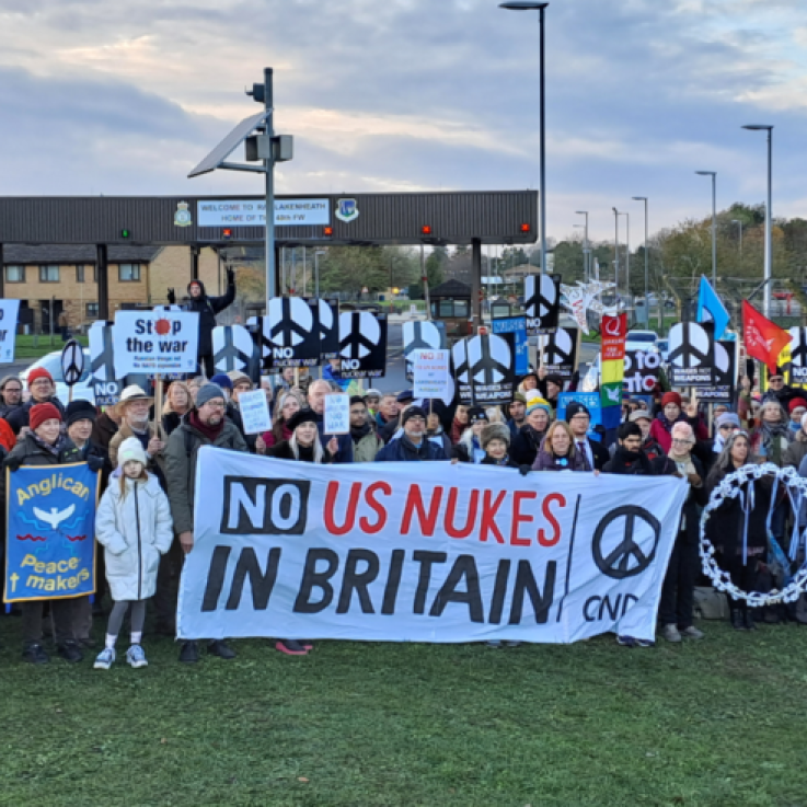 A large group of people stand outside, behind a banner reading "No US Nukes in Britain". In the background is a sign for RAF Lakenheath